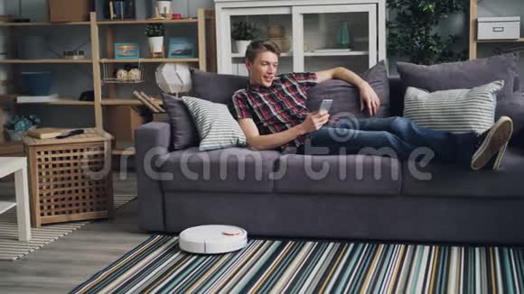 微笑的家伙正在上网智能手机在沙发上放松而机器人吸尘器正在清理地板视频的预览图