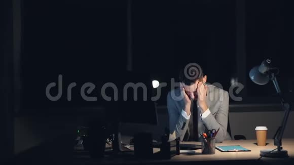 一个穿着西装的疲惫的年轻人正在电脑上工作深夜独自坐在办公室完成工作擦着脸视频的预览图