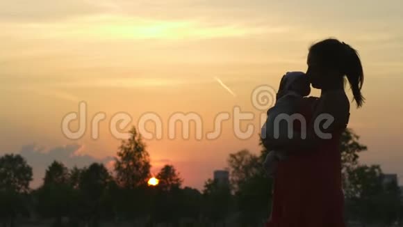 夕阳的剪影年轻的母亲抱着她的小男孩在城市公园站在夕阳面前生动活泼视频的预览图