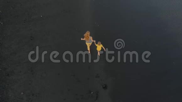 一位母亲和她的儿子在一片黑色火山沙的海滩上散步和玩耍的空中镜头视频的预览图