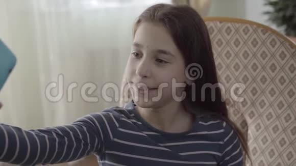 快乐可爱的十几岁女孩用她的蓝色手机自拍坐在家里的扶手椅上特写无忧无虑的孩子视频的预览图
