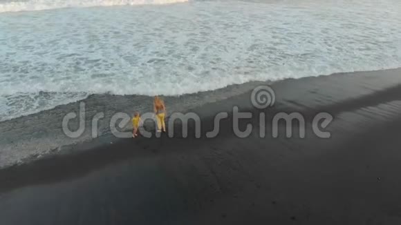 一位母亲和她的儿子在一片黑色火山沙的海滩上散步和玩耍的空中镜头视频的预览图