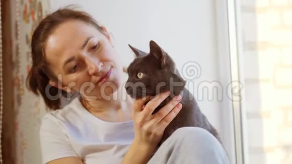 一个穿着白色T恤的女人正坐在窗台上戴着一只灰色的猫视频的预览图