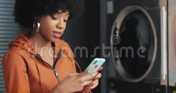 快乐迷人的非裔美国年轻女子在自助洗衣店智能手机上自拍自助公共洗衣店视频的预览图