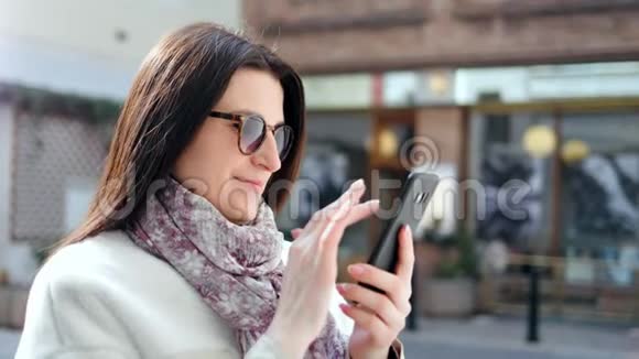 在现代城市街道上戴着墨镜用智能手机打字的迷人微笑女人视频的预览图