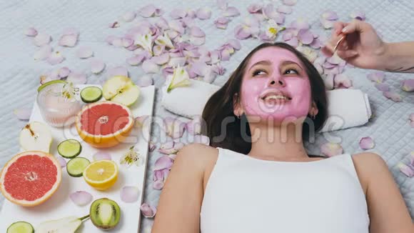 快乐的年轻女子在健康中心放松而美容师则涂上粉红色的面具视频的预览图