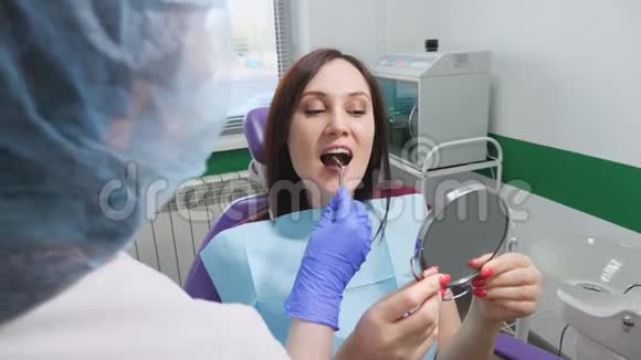 牙医正在给坐在牙椅上的年轻妇女看治疗后镜子里的牙齿视频的预览图