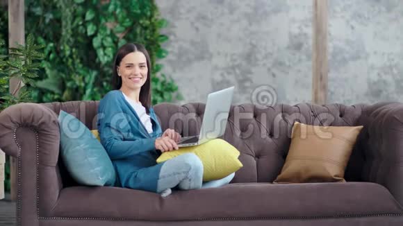 休闲微笑的女商人双腿交叉坐在沙发上享受周末视频的预览图