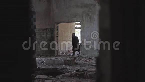 一个穿着黑色雨衣帽子和眼镜的神秘人穿过一家废弃工厂的破碎房间视频的预览图