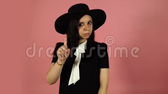 穿黑色连衣裙和帽子的年轻女性有魅力的女人穿着黑色的衣服站在粉红色的背景上指着视频的预览图