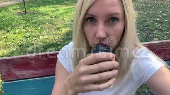 年轻的金发女郎坐在夏季城市公园的长凳上喝着咖啡一次性的纸带着一个带着外卖的杯子视频的预览图