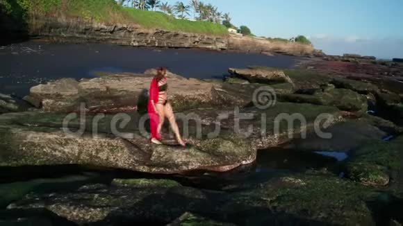 在黑色沙滩附近的岩石上漫步的美丽性感的比基尼女郎和红色的整体景观视频的预览图