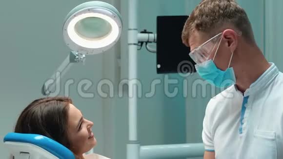 年轻英俊的牙医戴着蓝色的医用手套口罩和眼镜与深褐色皮肤的病人讨论女人的过程视频的预览图