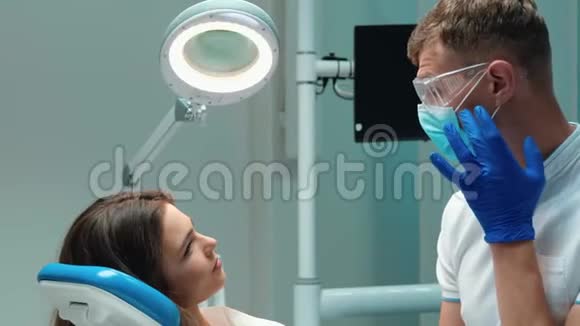年轻英俊的牙医戴着医用蓝色手套口罩和眼镜与美丽的病人讨论过程视频的预览图