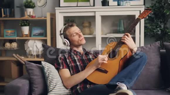 帅哥美男正在听耳机里的音乐弹吉他在家里的沙发上休闲放松视频的预览图