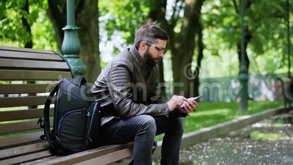 有胡子的人在长凳上的公园里用电话视频的预览图