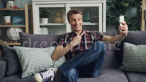 穿休闲服装的快乐男人正在用智能手机相机进行自拍玩得开心微笑坐在沙发上摆姿势视频的预览图