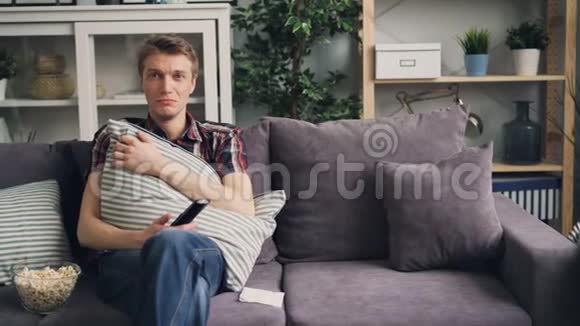 有魅力的男生学生在电视上为悲伤的戏剧哭泣年轻人坐在舒适的沙发上拿着遥控器视频的预览图