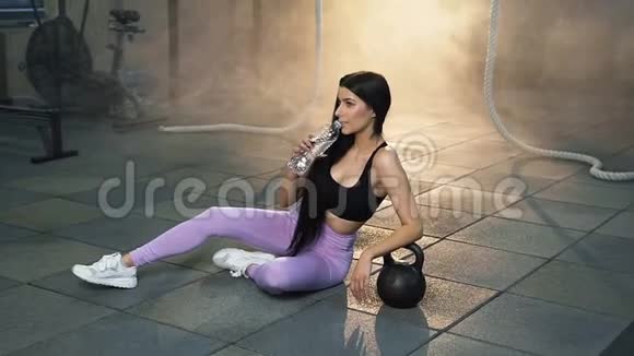 侧面看性感的年轻女人在健身房里躺在地板上喝水视频的预览图