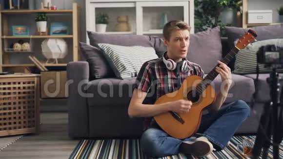 成功的年轻男子vlogger正在录制视频弹吉他坐在家里的地板上唱歌并使用相机视频的预览图