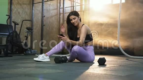 微笑运动女性在训练后休息并使用智能手机在健身房聊天视频的预览图
