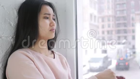 头痛沮丧的年轻亚洲女人坐在窗前视频的预览图
