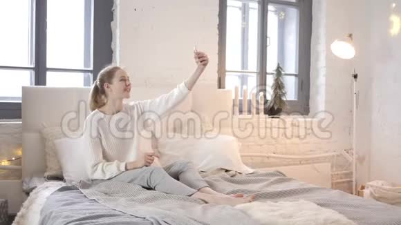 快乐女孩坐在床上用电话自拍照片视频的预览图