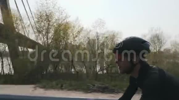 骑自行车的骑自行车者的特写镜头带着黑色头盔和墨镜骑着自行车骑着桥和河视频的预览图