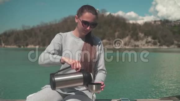 一个女人在阳光下喝热水瓶里的茶在春天的一天坐在湖边的一个木墩上在大自然中放松视频的预览图