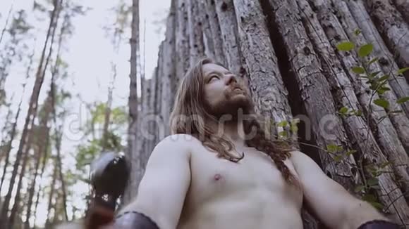 野蛮人坐在树林里靠在一棵树上拿着一把刀严肃地看着天空视频的预览图
