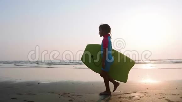 日落时小女孩正带着冲浪板沿着海边沙滩散步视频的预览图