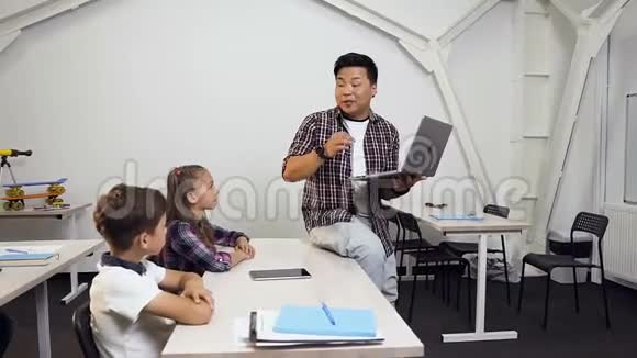 韩国男老师坐在桌子上手里拿着笔记本电脑给学生上课学龄儿童认真倾听视频的预览图