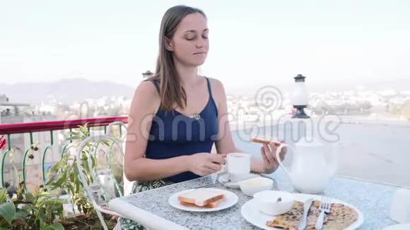 女人正在城市和山地背景的露台上的咖啡馆里吃早餐视频的预览图