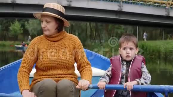 小可爱的孩子穿着救生衣和他的祖母坐在湖边的木船上航行可爱的男孩和孙子在船上漂浮视频的预览图