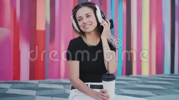 年轻女子一边喝着咖啡一边听着音乐一边坐在路边背景是一色的墙视频的预览图