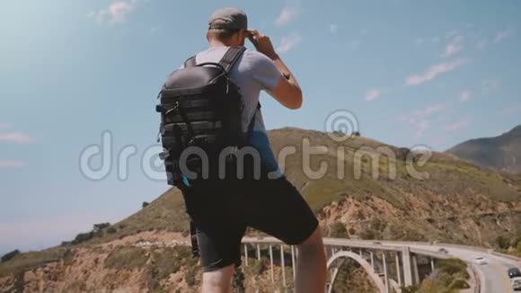 年轻的快乐旅游摄影师带着专业背包在加州比克斯比峡谷大桥观看史诗般的风景视频的预览图