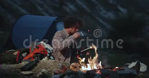 有魅力的卷发年轻人在帐篷旁露营坐在篝火旁喝着茶手里拿着一杯茶视频的预览图