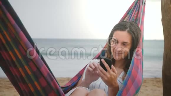 快乐成功的年轻女商人在海滩吊床在惊人的海洋海岸度假村使用智能手机移动办公应用程序视频的预览图