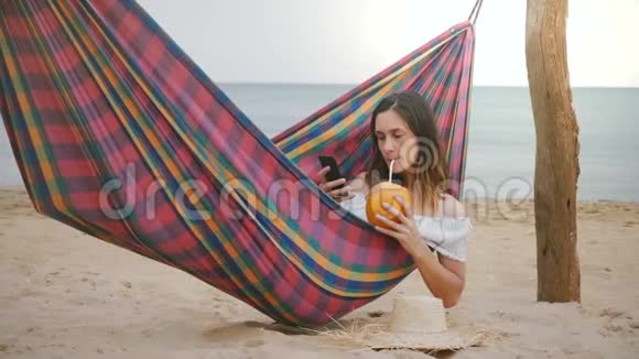 快乐成功的年轻女商人在海滨海滩吊床与水果鸡尾酒使用智能手机移动游戏应用程序视频的预览图