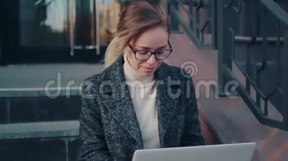 戴眼镜的年轻女作家在户外使用笔记本电脑穿外套的女孩坐在台阶上视频的预览图