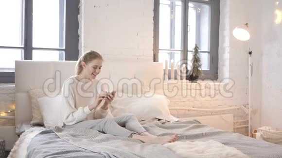 兴奋的女孩坐在床上庆祝智能手机的成功视频的预览图