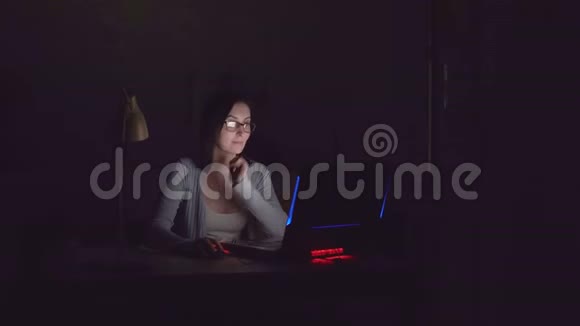 可爱的年轻女子在黑暗中微笑着看着家里的笔记本电脑屏幕视频的预览图