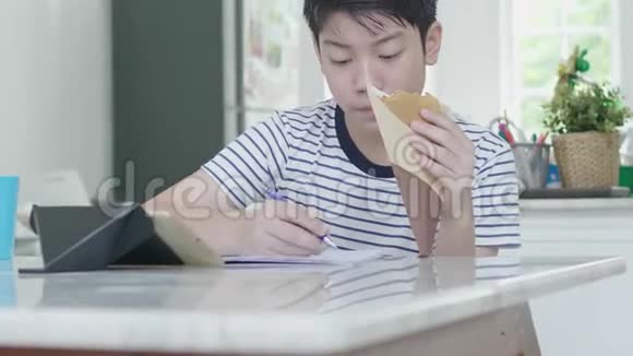 一个亚洲男孩在家做作业表情愉快手里拿着食物视频的预览图