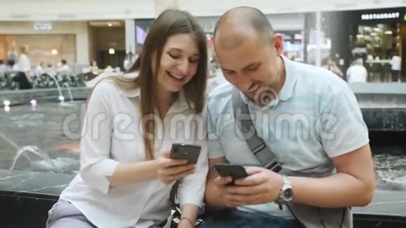 可爱的夫妇坐在购物中心的喷泉附近聊天看着手机上的照片视频的预览图