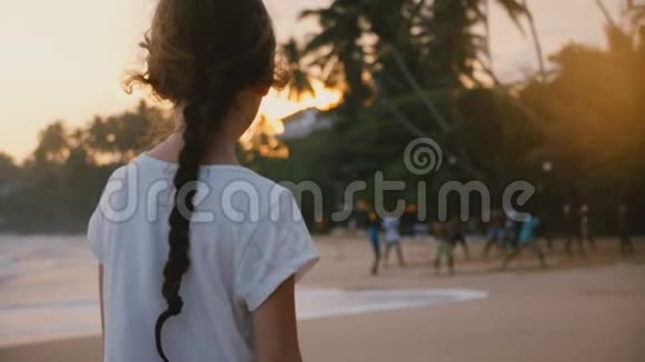 令人惊叹的后景拍摄小快乐女孩在热带异国情调的日落海滩观看当地人打排球视频的预览图