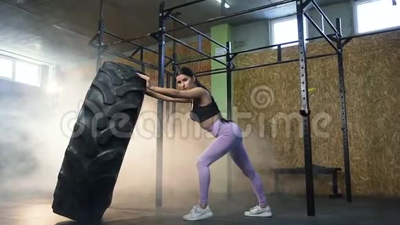 强壮的年轻女人拿着大拖拉机轮胎在健身房交叉训练时看着相机视频的预览图