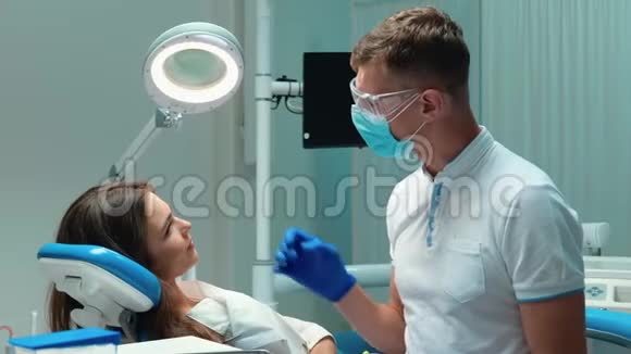 年轻英俊的牙医戴着医用蓝色手套口罩和眼镜与美丽的病人讨论过程视频的预览图