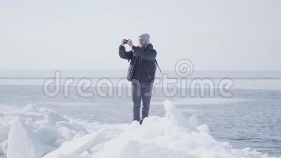 穿着温暖的冬衣戴着眼镜的英俊的金发大胡子男子站在冰川的冰块上拍照视频的预览图