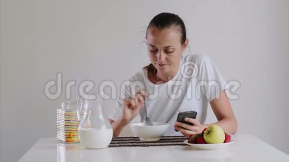 年轻的女人正在吃早餐看着智能手机视频的预览图