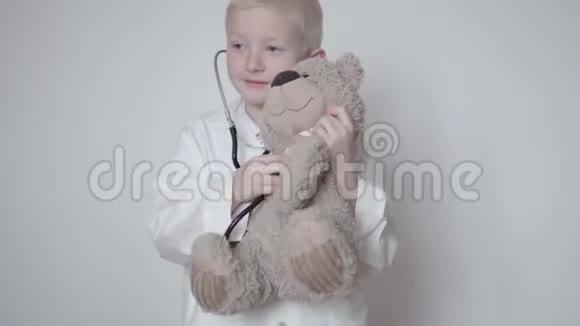 穿着医生外衣可爱的男孩在听一只带听诊器的玩具熊视频的预览图